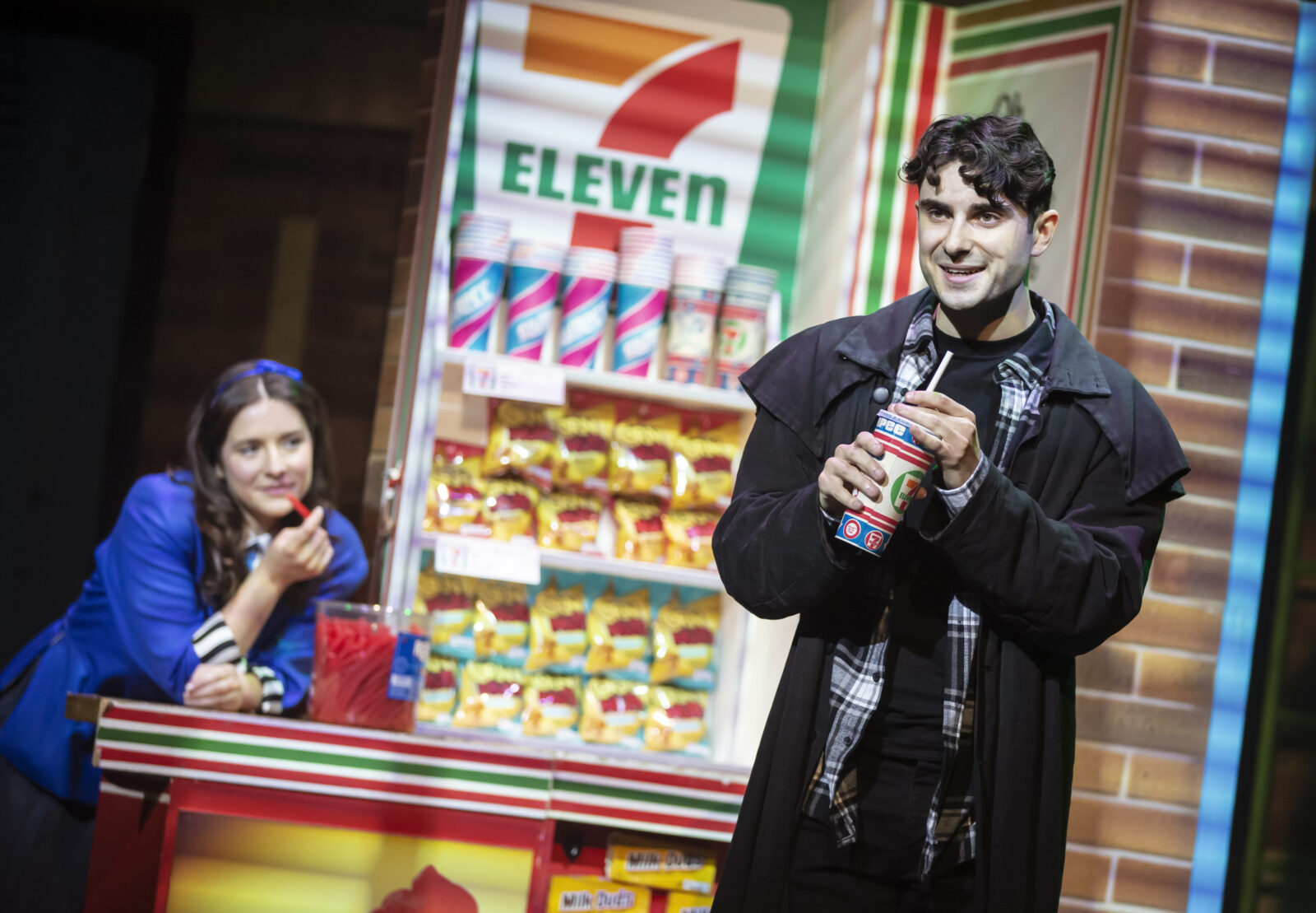 A man in a black coat onstage drinking a milkshake. 