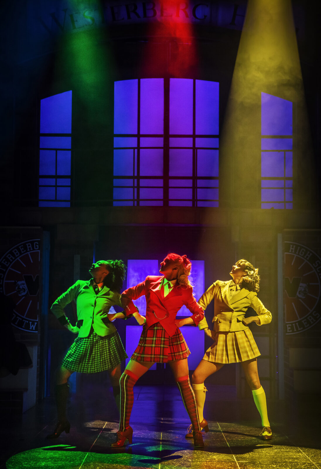 Three girls on stage with their hands on their hips wearing green, red and yellow school uniform. 
